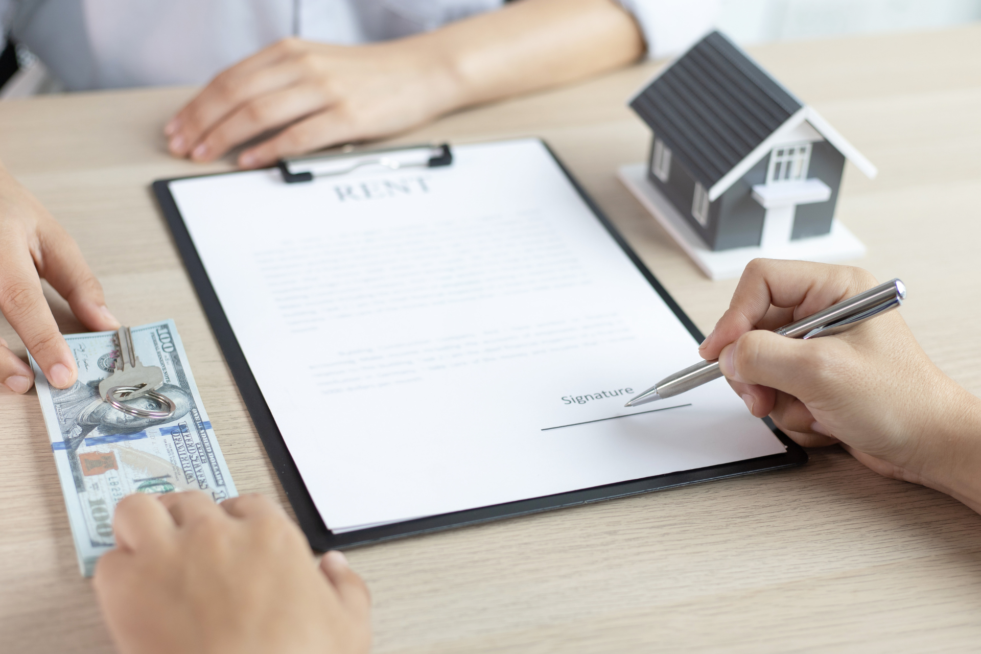 Two people sitting at a desk. One person is about to sign rental paperwork and they are both exchanging cash and a house key. There is a small fake house on the desk.