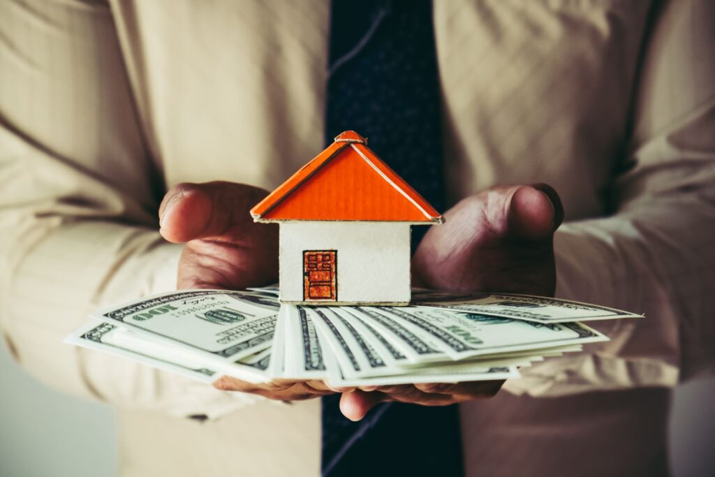 Hands of a man in a beige blazer holding out dollars and a miniature house with an orange door and roof, symbolizing the no security deposit option.