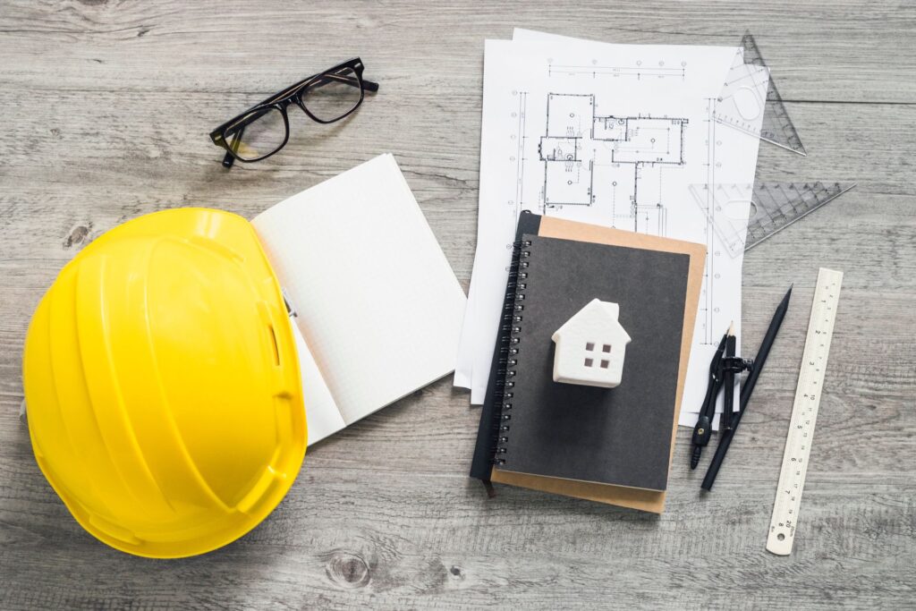 A gray table displaying a yellow hard hat, black glasses, a ruler, a pen, blueprints, and a black notebook, with a small model house placed on top of the notebook, representing new construction builds.