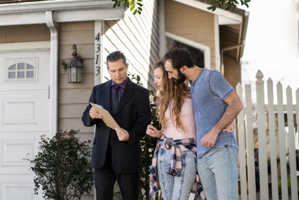 A realtor reviews paperwork with a young couple outside a house, indicating a positive experience for first-time home buyers.