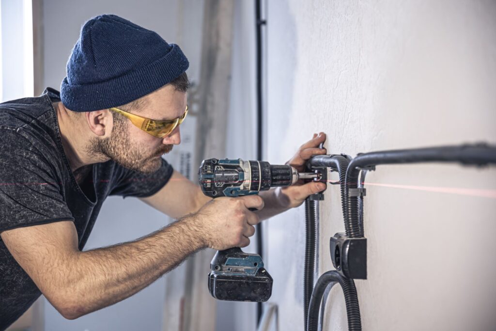 A man wearing safety glasses and a beanie uses a drill on a white wall with black cables attached. A red laser line is visible, illustrating tenant tips to prevent maintenance issues.