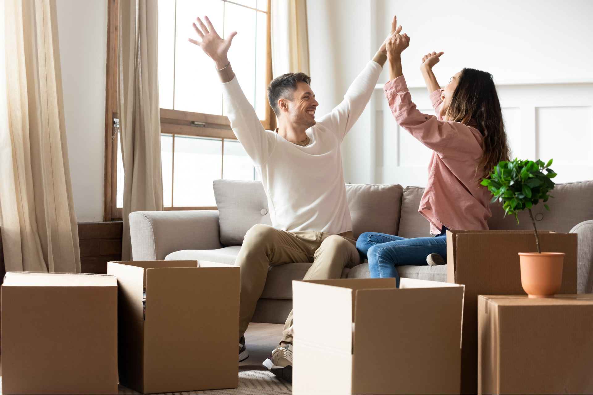 A joyful couple sitting on a sofa in their new home, surrounded by unpacked moving boxes. They are celebrating their move with arms raised, expressing excitement and accomplishment. This scene illustrates how to improve your Orlando rental experience.